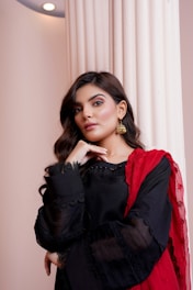A woman with long, wavy hair wears a black traditional outfit with intricate lace detailing and a vibrant red scarf draped over her shoulder. She accessorizes with gold earrings and stands next to a white column, posing with her chin resting on her hand.