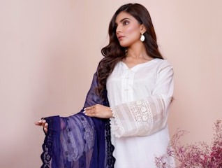 A woman is wearing a white dress with intricate lace details on the sleeves and holding a dark blue embroidered shawl. Her hair is styled in loose waves, and she is wearing drop earrings. The background is a soft beige color, and there are delicate pink flowers on the lower right side.