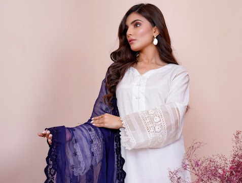 A woman is wearing a white dress with intricate lace details on the sleeves and holding a dark blue embroidered shawl. Her hair is styled in loose waves, and she is wearing drop earrings. The background is a soft beige color, and there are delicate pink flowers on the lower right side.