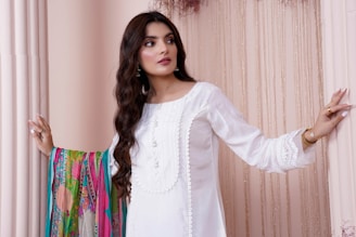 A woman with long, wavy dark hair stands gracefully in a white embroidered dress. She is positioned between light pink columns and holds a vibrantly colored shawl. The background features delicate, flowing curtains.