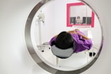 A person is lying down inside a medical imaging device, such as an MRI or CT scanner. The scanner surrounds the individual, who is wearing a purple garment. In the background, there is a control room with a glass window where a person is visible operating the equipment.