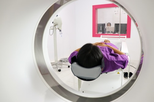 A person is lying down inside a medical imaging device, such as an MRI or CT scanner. The scanner surrounds the individual, who is wearing a purple garment. In the background, there is a control room with a glass window where a person is visible operating the equipment.