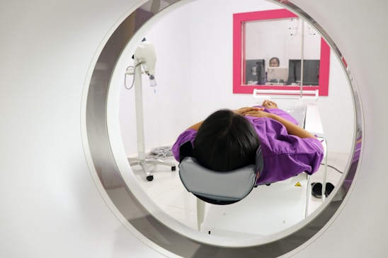 A person is lying down inside a medical imaging device, such as an MRI or CT scanner. The scanner surrounds the individual, who is wearing a purple garment. In the background, there is a control room with a glass window where a person is visible operating the equipment.