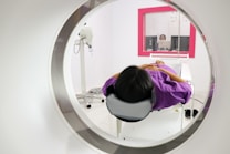 A person is lying down inside a medical imaging device, such as an MRI or CT scanner. The scanner surrounds the individual, who is wearing a purple garment. In the background, there is a control room with a glass window where a person is visible operating the equipment.