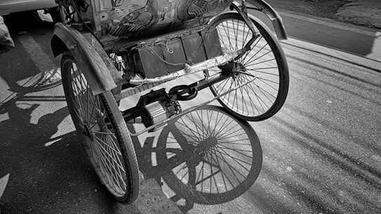 A close-up view of a rickshaw wheel casting a shadow on the street. The wheel's intricate spokes create a detailed pattern on the ground. The back of the rickshaw, featuring decorative elements, is partially visible. The image is in black and white, enhancing the play of light and shadow.