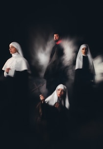 A group of individuals wearing religious habits are positioned in a dimly lit setting, evoking a sense of mystery and solemnity. One holds a cross, gazing upward, while another looks contemplatively to the side. The others stand or sit in a ghostly mist, adding a surreal quality.