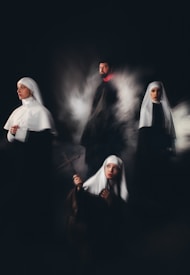 A group of individuals wearing religious habits are positioned in a dimly lit setting, evoking a sense of mystery and solemnity. One holds a cross, gazing upward, while another looks contemplatively to the side. The others stand or sit in a ghostly mist, adding a surreal quality.