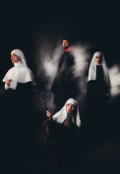 A group of individuals wearing religious habits are positioned in a dimly lit setting, evoking a sense of mystery and solemnity. One holds a cross, gazing upward, while another looks contemplatively to the side. The others stand or sit in a ghostly mist, adding a surreal quality.