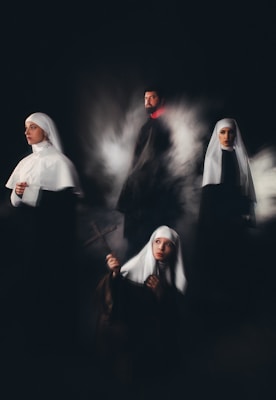 A group of individuals wearing religious habits are positioned in a dimly lit setting, evoking a sense of mystery and solemnity. One holds a cross, gazing upward, while another looks contemplatively to the side. The others stand or sit in a ghostly mist, adding a surreal quality.
