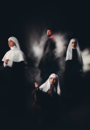A group of individuals wearing religious habits are positioned in a dimly lit setting, evoking a sense of mystery and solemnity. One holds a cross, gazing upward, while another looks contemplatively to the side. The others stand or sit in a ghostly mist, adding a surreal quality.