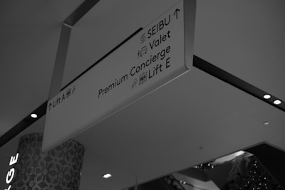 A monochrome photograph of a ceiling-mounted directional sign in a commercial or public space. The sign displays directions to various services including SEIBU, valet, premium concierge, and lifts A and E. The background shows part of a building interior with patterned walls and ceiling lights.