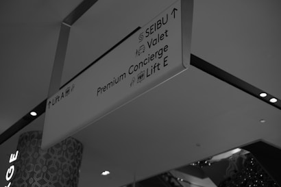 A monochrome photograph of a ceiling-mounted directional sign in a commercial or public space. The sign displays directions to various services including SEIBU, valet, premium concierge, and lifts A and E. The background shows part of a building interior with patterned walls and ceiling lights.
