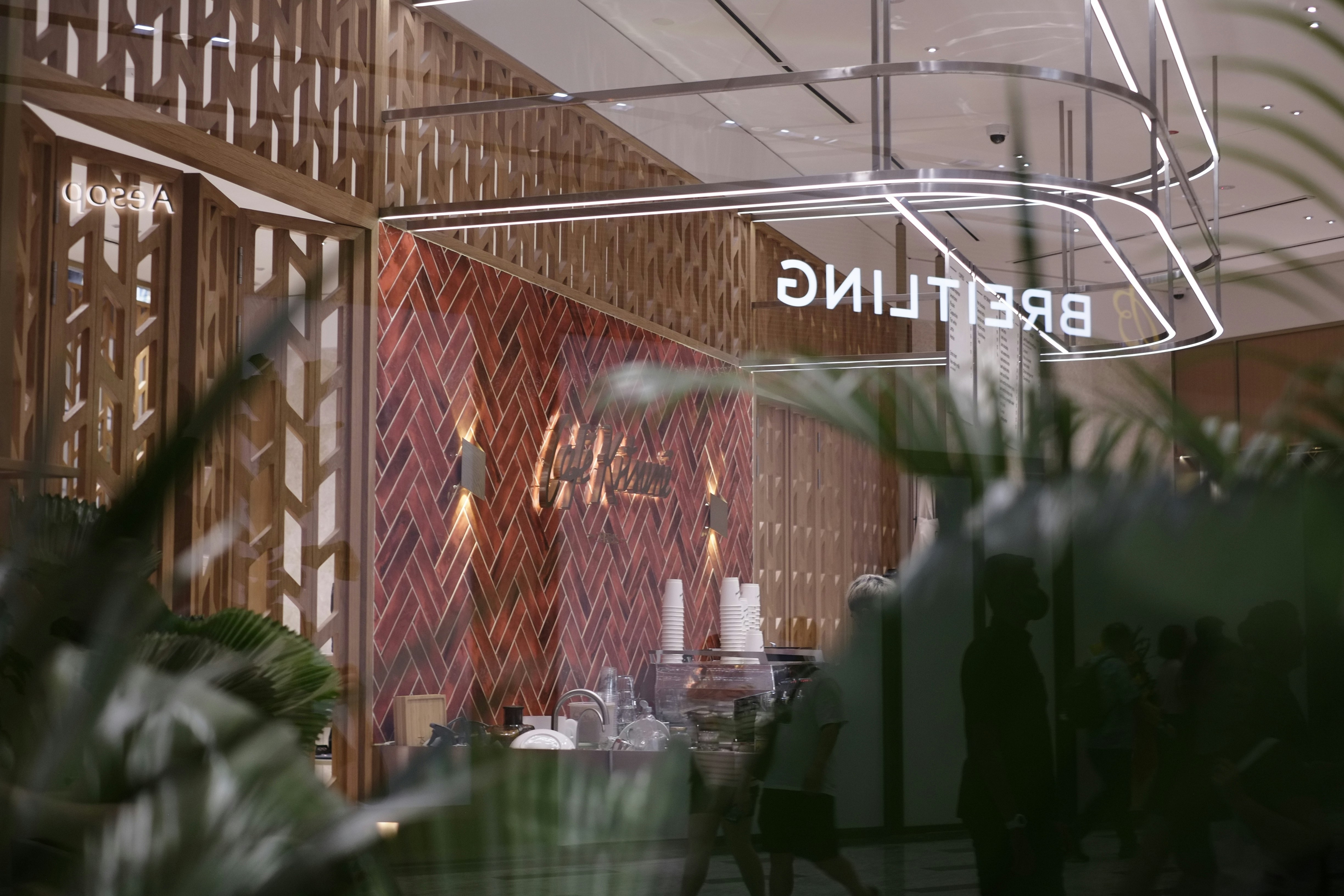 A modern retail space with a sophisticated design, featuring a herringbone-patterned wall made of warm wood tones. The reflection shows the store's name, 'BREITLING,' in bright, illuminated letters. The interior includes various items like stacked cups and other objects on a counter. Large glass windows create a sense of openness, with greenery visible in the foreground, adding a touch of nature to the scene.