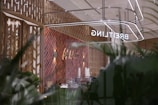 A modern retail space with a sophisticated design, featuring a herringbone-patterned wall made of warm wood tones. The reflection shows the store's name, 'BREITLING,' in bright, illuminated letters. The interior includes various items like stacked cups and other objects on a counter. Large glass windows create a sense of openness, with greenery visible in the foreground, adding a touch of nature to the scene.