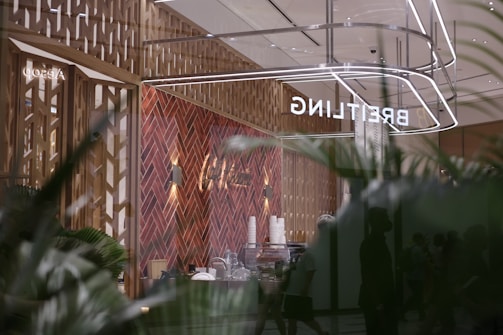 A modern retail space with a sophisticated design, featuring a herringbone-patterned wall made of warm wood tones. The reflection shows the store's name, 'BREITLING,' in bright, illuminated letters. The interior includes various items like stacked cups and other objects on a counter. Large glass windows create a sense of openness, with greenery visible in the foreground, adding a touch of nature to the scene.