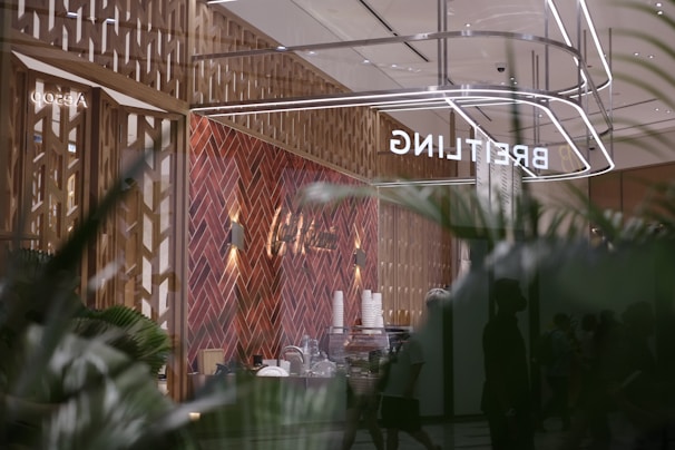 A modern retail space with a sophisticated design, featuring a herringbone-patterned wall made of warm wood tones. The reflection shows the store's name, 'BREITLING,' in bright, illuminated letters. The interior includes various items like stacked cups and other objects on a counter. Large glass windows create a sense of openness, with greenery visible in the foreground, adding a touch of nature to the scene.