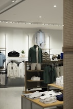 A retail clothing store display featuring mannequins dressed in casual, sporty attire including jackets and shorts. Shelves and racks are organized with neatly folded shirts and jackets in neutral and earthy tones. The lighting is bright and modern, enhancing the sleek display of garments and accessories.