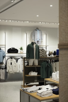 A retail clothing store display featuring mannequins dressed in casual, sporty attire including jackets and shorts. Shelves and racks are organized with neatly folded shirts and jackets in neutral and earthy tones. The lighting is bright and modern, enhancing the sleek display of garments and accessories.