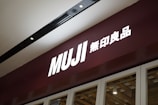 A storefront sign with the brand name Muji, featuring large white letters on a dark maroon background. The sign includes both English and Japanese characters. The lighting is focused with a sleek and modern ceiling fixture visible above.