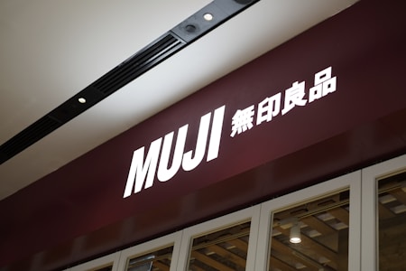A storefront sign with the brand name Muji, featuring large white letters on a dark maroon background. The sign includes both English and Japanese characters. The lighting is focused with a sleek and modern ceiling fixture visible above.