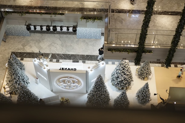 An elegantly designed mall interior features a large display area adorned with several artificial, white-frosted Christmas trees. A prominent luxury brand logo is placed centrally against a well-lit display wall. The floor is marble, and decorative garlands are hung along the railings and edges. The environment conveys a festive, upscale shopping atmosphere.