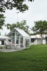 A small, modern glass house is situated in a lush green garden, surrounded by trees. There are several chairs and tables outside under a large umbrella, suggesting a cozy seating or dining area. The architecture is mostly minimalistic with an emphasis on glass, creating a transparent and airy look.
