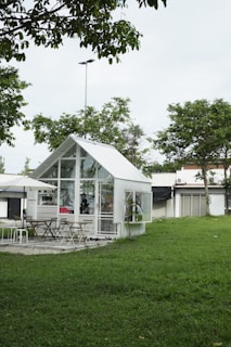A small, modern glass house is situated in a lush green garden, surrounded by trees. There are several chairs and tables outside under a large umbrella, suggesting a cozy seating or dining area. The architecture is mostly minimalistic with an emphasis on glass, creating a transparent and airy look.