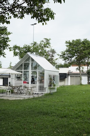 A small, modern glass house is situated in a lush green garden, surrounded by trees. There are several chairs and tables outside under a large umbrella, suggesting a cozy seating or dining area. The architecture is mostly minimalistic with an emphasis on glass, creating a transparent and airy look.
