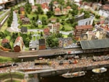 A detailed miniature diorama depicting a bustling town with a railway line running through it. The scene includes a train station, numerous intricately designed buildings, trees, and winding roads. The landscape is lush and green, with a variety of architectural styles in the buildings, giving it a quaint, small-town charm.