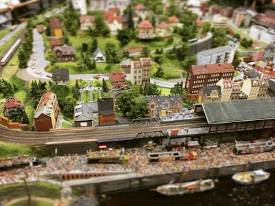 A detailed miniature diorama depicting a bustling town with a railway line running through it. The scene includes a train station, numerous intricately designed buildings, trees, and winding roads. The landscape is lush and green, with a variety of architectural styles in the buildings, giving it a quaint, small-town charm.