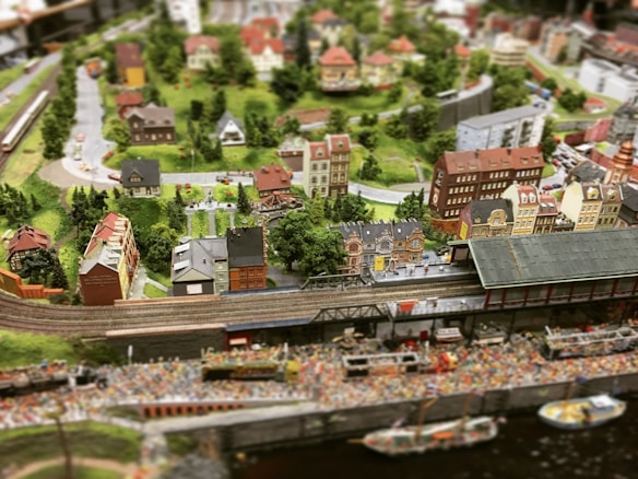 A detailed miniature diorama depicting a bustling town with a railway line running through it. The scene includes a train station, numerous intricately designed buildings, trees, and winding roads. The landscape is lush and green, with a variety of architectural styles in the buildings, giving it a quaint, small-town charm.