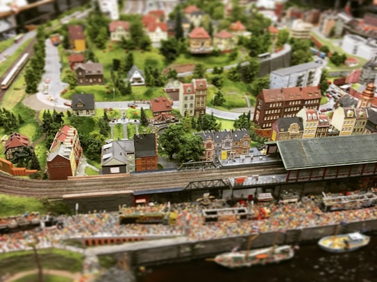 A detailed miniature diorama depicting a bustling town with a railway line running through it. The scene includes a train station, numerous intricately designed buildings, trees, and winding roads. The landscape is lush and green, with a variety of architectural styles in the buildings, giving it a quaint, small-town charm.
