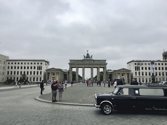 A wide stone plaza leads up to a large neoclassical monument known as the Brandenburg Gate, surrounded by tourists. The structure features columns and a chariot sculpture atop its summit. To the sides, two symmetrical buildings frame the scene. A stretch limousine is parked in the foreground on the right.