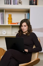 A woman with wavy brown hair is seated in a wooden chair using a black laptop. She is dressed in a dark top and pants. Behind her is a white shelving unit holding various books, a yellow vase, a stack of paper, and a small white clock.