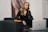 A woman with long blonde hair and glasses is sitting on a dark gray couch. She is wearing a black dress and is leaning toward a laptop on a dark table. There is a bright orange book placed on the arm of the couch, and a modern abstract painting hangs on the wall behind her.