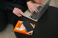A person is typing on a laptop while sitting next to a black table. On the table, there is an orange book and a smartphone placed on top of it. The individual is wearing a black outfit and has black nail polish.