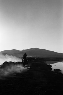 A silhouetted cowboy rides on horseback along a dusty trail with mountains in the background and a body of water on the right. Dust rises from the ground, creating a hazy atmosphere.