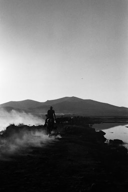 A silhouetted cowboy rides on horseback along a dusty trail with mountains in the background and a body of water on the right. Dust rises from the ground, creating a hazy atmosphere.
