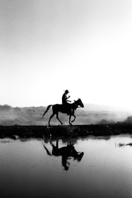 A silhouette of a person riding a horse along a path with a reflective surface in the foreground, possibly water. The background features a clear sky with gentle gradients, and the overall setting seems tranquil with natural elements like distant hills or mountains.