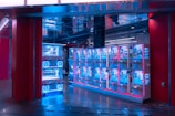 A row of brightly lit claw machines stands inside a modern arcade. The machines have a glowing blue hue, accented by pink and red lighting. The area is clean, with a shiny floor reflecting the neon lights, and there's a ceiling with mirrored panels creating a visually intriguing atmosphere.