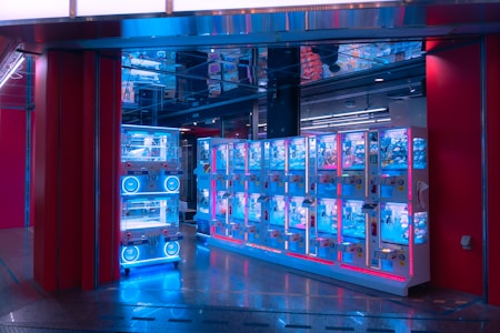 A row of brightly lit claw machines stands inside a modern arcade. The machines have a glowing blue hue, accented by pink and red lighting. The area is clean, with a shiny floor reflecting the neon lights, and there's a ceiling with mirrored panels creating a visually intriguing atmosphere.