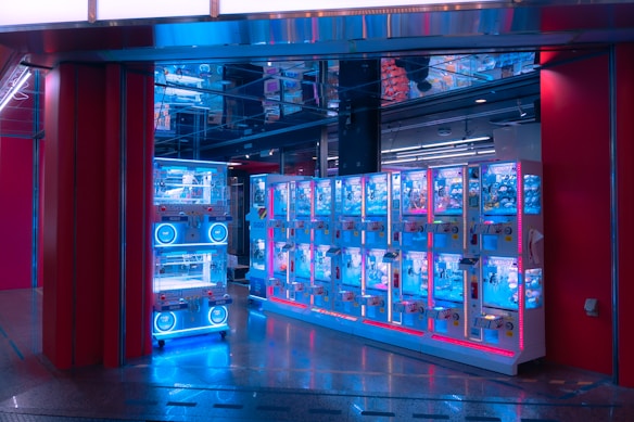 A row of brightly lit claw machines stands inside a modern arcade. The machines have a glowing blue hue, accented by pink and red lighting. The area is clean, with a shiny floor reflecting the neon lights, and there's a ceiling with mirrored panels creating a visually intriguing atmosphere.