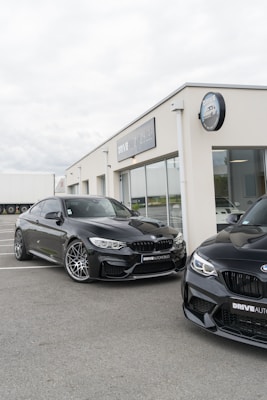 Two black sports cars are parked in front of a modern auto dealership with large glass windows and a sign displaying 'DRIVE AUTOMOBILES'. The overcast sky creates a neutral backdrop for the sleek vehicles.