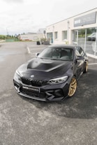 A sleek black BMW car with gold wheels is parked in front of an automotive dealership. The dealership has large glass windows and the name 'DRIVE AUTOMOBILES' is displayed on the building. The surroundings include paved ground and some vegetation in the background.
