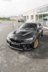 A sleek black BMW car with gold wheels is parked in front of an automotive dealership. The dealership has large glass windows and the name 'DRIVE AUTOMOBILES' is displayed on the building. The surroundings include paved ground and some vegetation in the background.