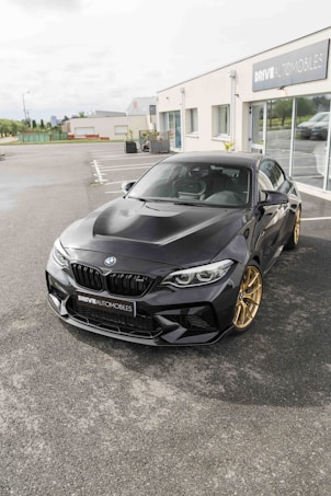 A sleek black BMW car with gold wheels is parked in front of an automotive dealership. The dealership has large glass windows and the name 'DRIVE AUTOMOBILES' is displayed on the building. The surroundings include paved ground and some vegetation in the background.