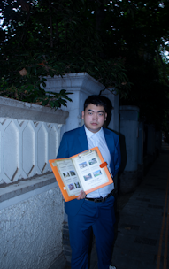 A person in a blue suit stands on a dimly lit pathway holding an orange folder open, displaying several photos and documents. The scene is framed by a stone wall and dense foliage.