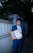 A person in a blue suit stands on a dimly lit pathway holding an orange folder open, displaying several photos and documents. The scene is framed by a stone wall and dense foliage.