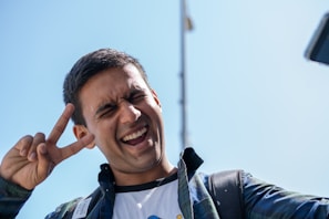 A person smiling broadly while making a peace sign with their hand. The individual is wearing a casual plaid shirt over a t-shirt and carries a backpack. The clear blue sky serves as the background.