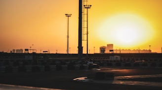 A large sun sets on the horizon, casting an orange glow over a go-kart track. Tall floodlights tower over the scene, with scattered structures in the distance. The track is outlined by tire barriers, and a lone go-kart can be seen near the center.